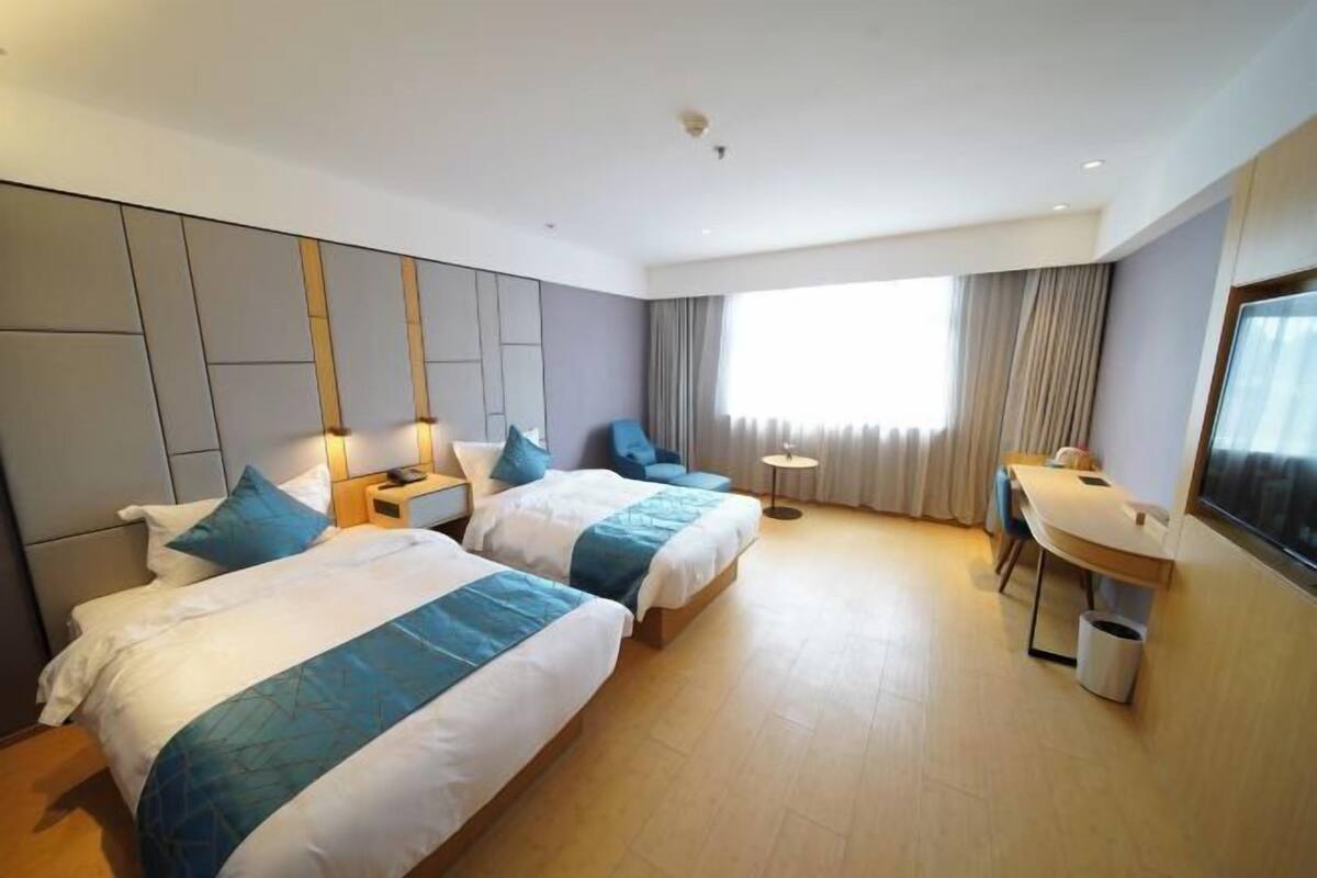 Ein Hotelzimmer mit zwei Betten, einem Schreibtisch, einem Stuhl, einem kleinen Tisch und einem großen Fenster mit Vorhängen.