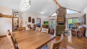 A spacious living area with a wooden dining table, comfortable seating, a stone fireplace, and large windows offering a view of the outdoors. - Newly Renovated Condo in Gated Golf Community (Whittier)