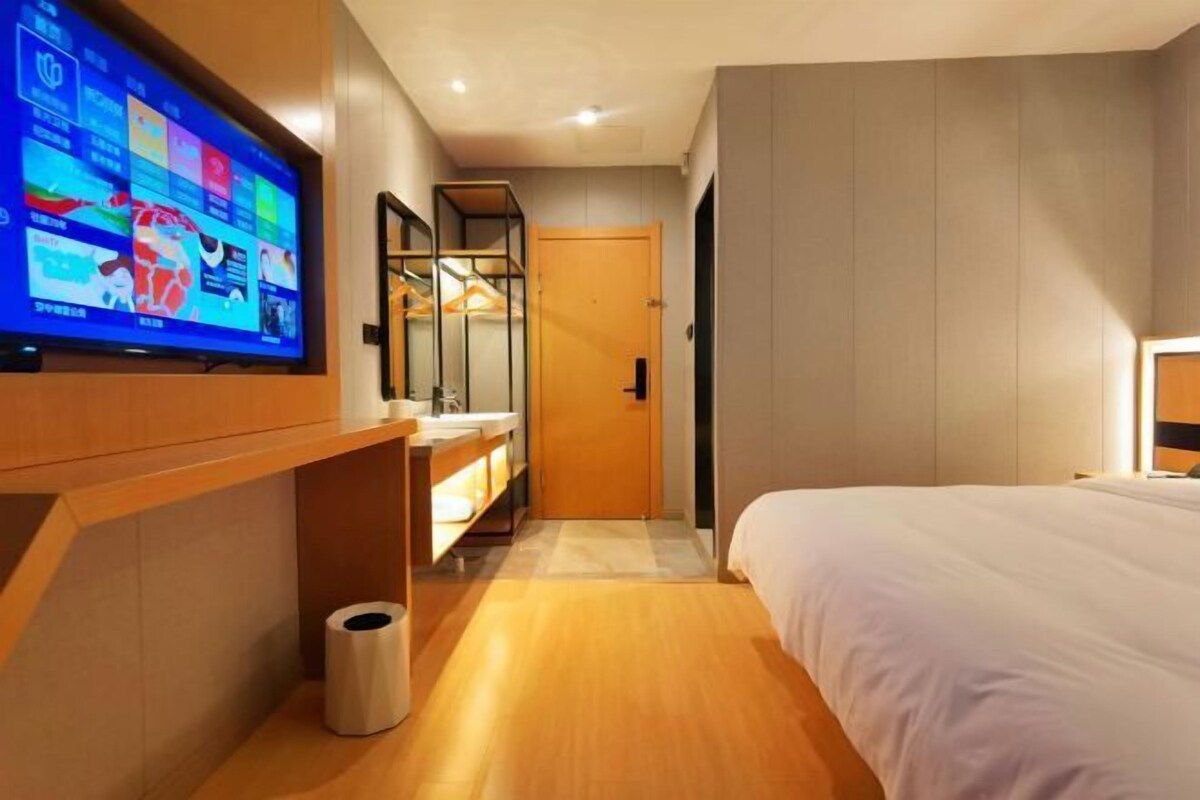 A modern hotel room with a large flat-screen TV mounted on the wall, a wooden vanity with a mirror, a bed with white bedding, and a wooden floor.