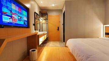 A modern hotel room with a large flat-screen TV mounted on the wall, a wooden vanity with a mirror, a bed with white bedding, and a wooden floor.