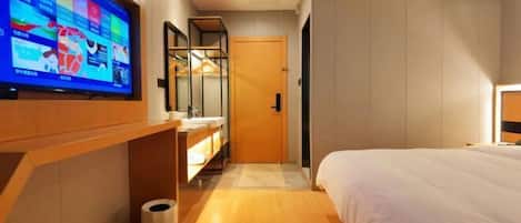 A modern hotel room with a large flat-screen TV mounted on the wall, a wooden vanity with a mirror, a bed with white bedding, and a wooden floor.