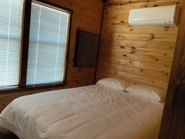 Een kamer met houten lambrisering, een bed, een raam met jaloezieën en een aan de muur bevestigde airconditioning.