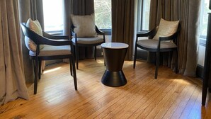 Una habitación moderna con suelo de madera, tres sillas oscuras con cojines, una pequeña mesa redonda y ventanas amplias con cortinas.