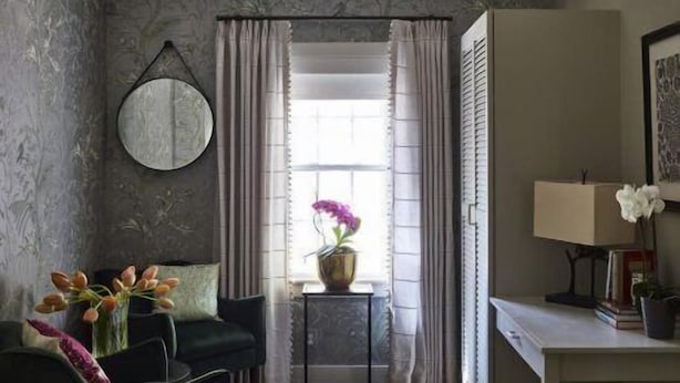 Ein Zimmer mit einem dunkelgrünen Sofa, einem kleinen Tisch mit Blumen, einem Schreibtisch mit einer Lampe und einem Fenster mit Vorhängen.