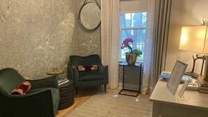 Ein Wohnzimmer mit einem grünen Sessel, einem runden Beistelltisch, einem kleinen Teppich, einem Schreibtisch mit Lampe und einem Fenster mit Vorhängen.
