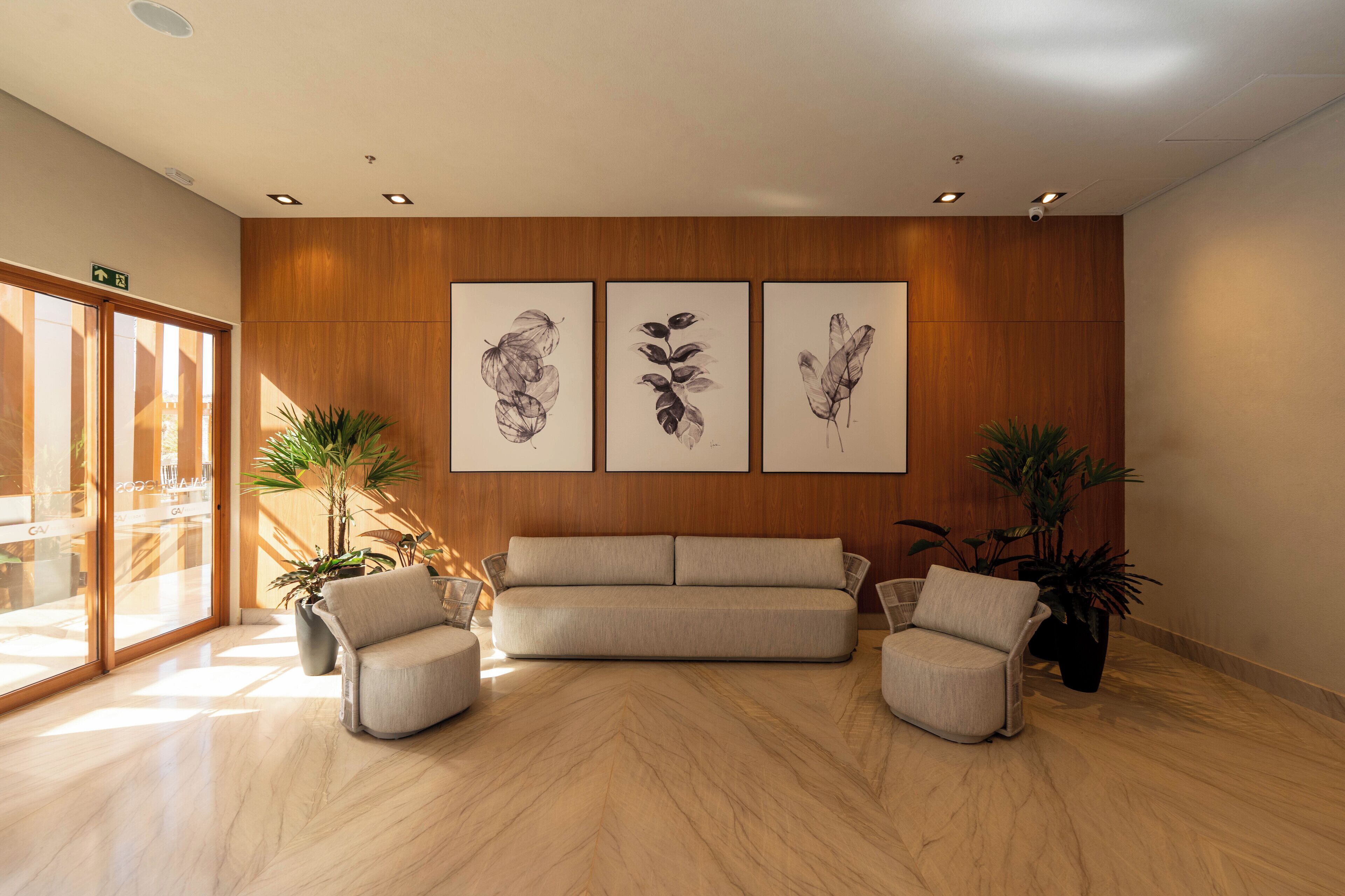 A modern lobby with a wooden wall, a beige sofa, two armchairs, and three framed botanical prints.