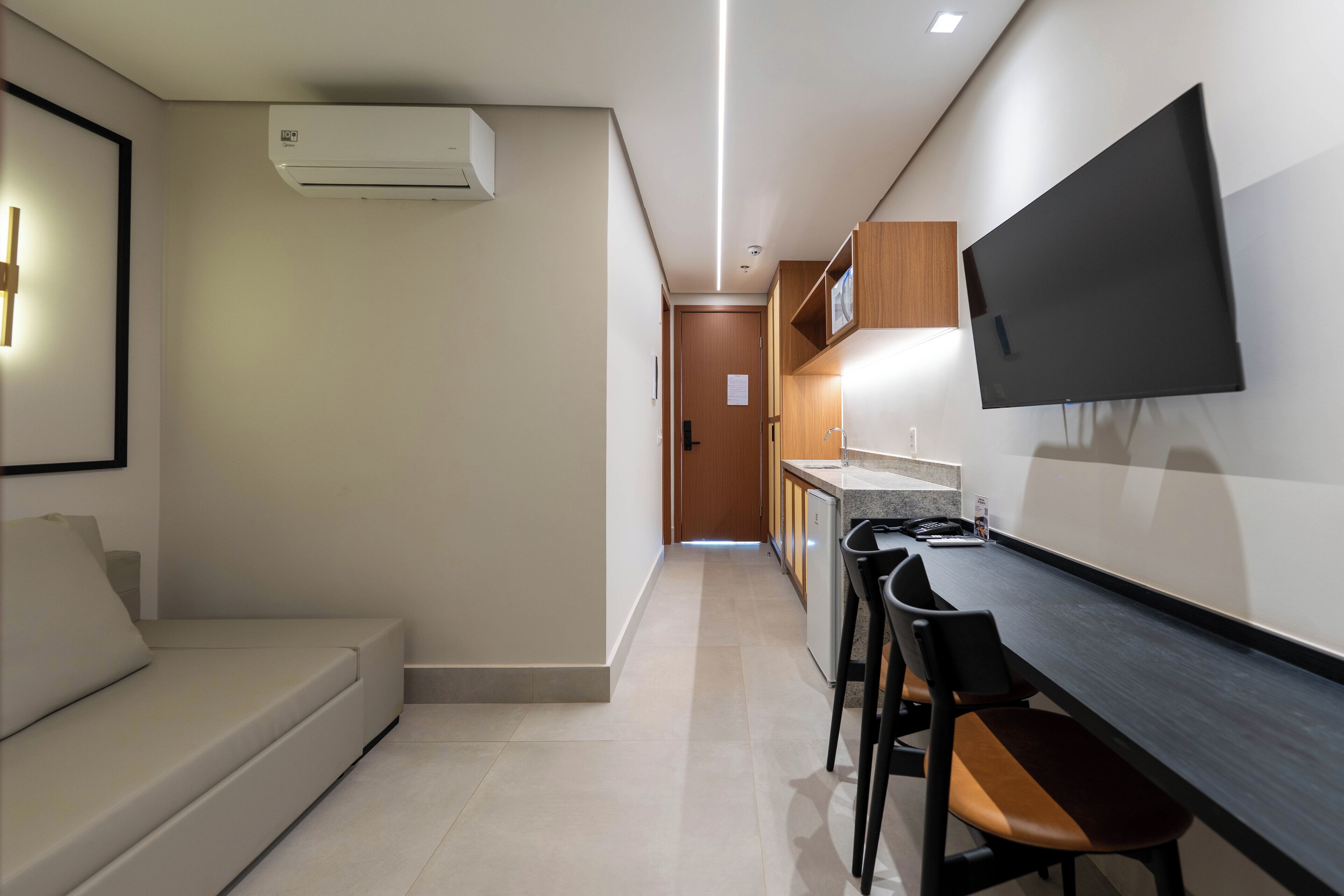A modern kitchen with a flat-screen TV mounted on the wall, a dining area with a long table and chairs, and a sofa in the adjacent living area.