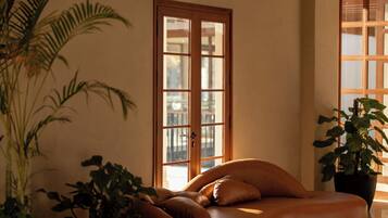 A room with a wooden ceiling, a large window, and a sofa with cushions. There are potted plants on the floor.