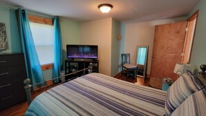 A bedroom with a bed, a television on a stand, a chair, a wooden door, and a window with curtains. - Charming 1-bedroom house on a lavender farm (Scarbro)