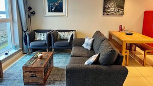 En moderne stue med sofa, to lænestole, et træ sofabord og et spisebord. To indrammede kunstværker hænger på væggen, og et vindue er dækket af gardiner.
