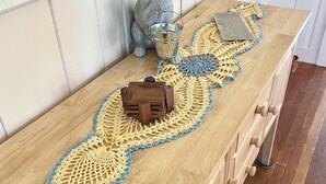 Una mesa de madera con un camino tejido al crochet, un cubo de metal y una maceta de cerámica.
