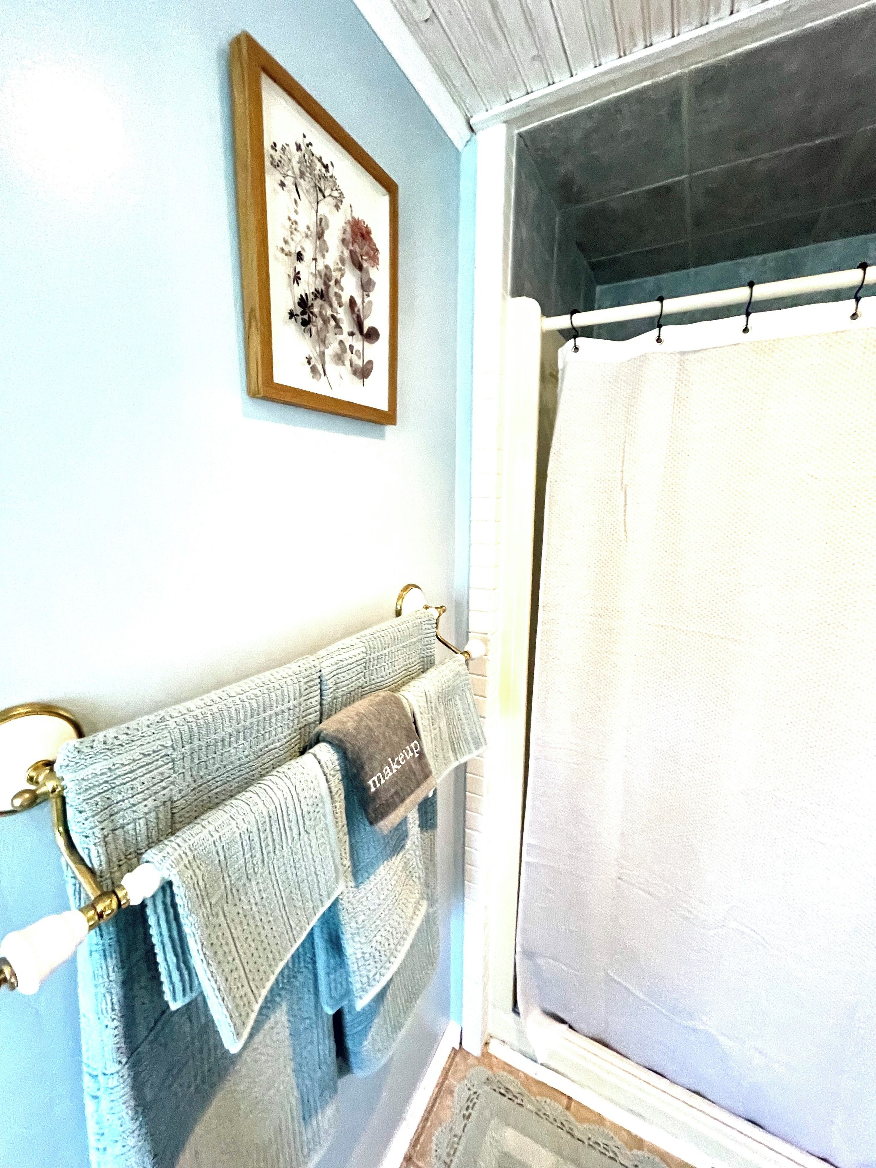 A bathroom with a shower curtain, a towel rack with towels, and a framed floral artwork on the wall.