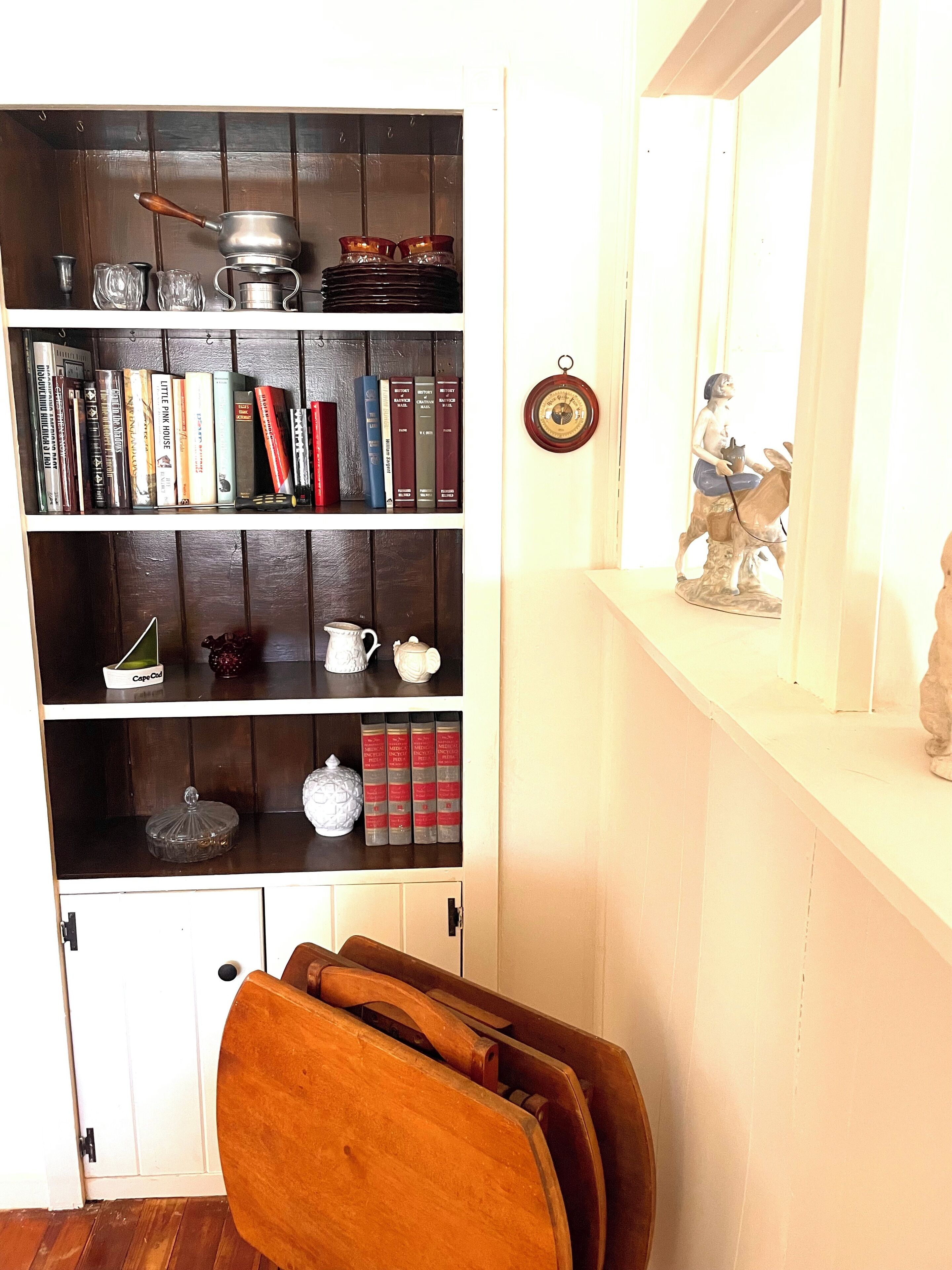 A room with a wooden bookshelf filled with books and decorative items, a white door, and a wooden chair.
