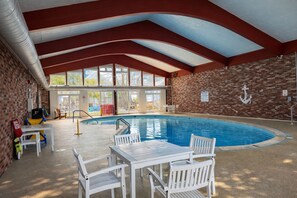 An indoor swimming pool with a brick wall, a white table with chairs, and a large window. - Courtyard Resort (Hyannis)