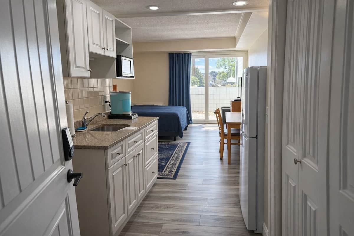 A compact hotel room with a kitchenette, a bed, a dining area, and a view through a large window.