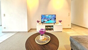 Ett modernt vardagsrum med en platt-TV placerad på en vit TV-bänk, ett runt soffbord med en vävd underlägg och en grå soffa.