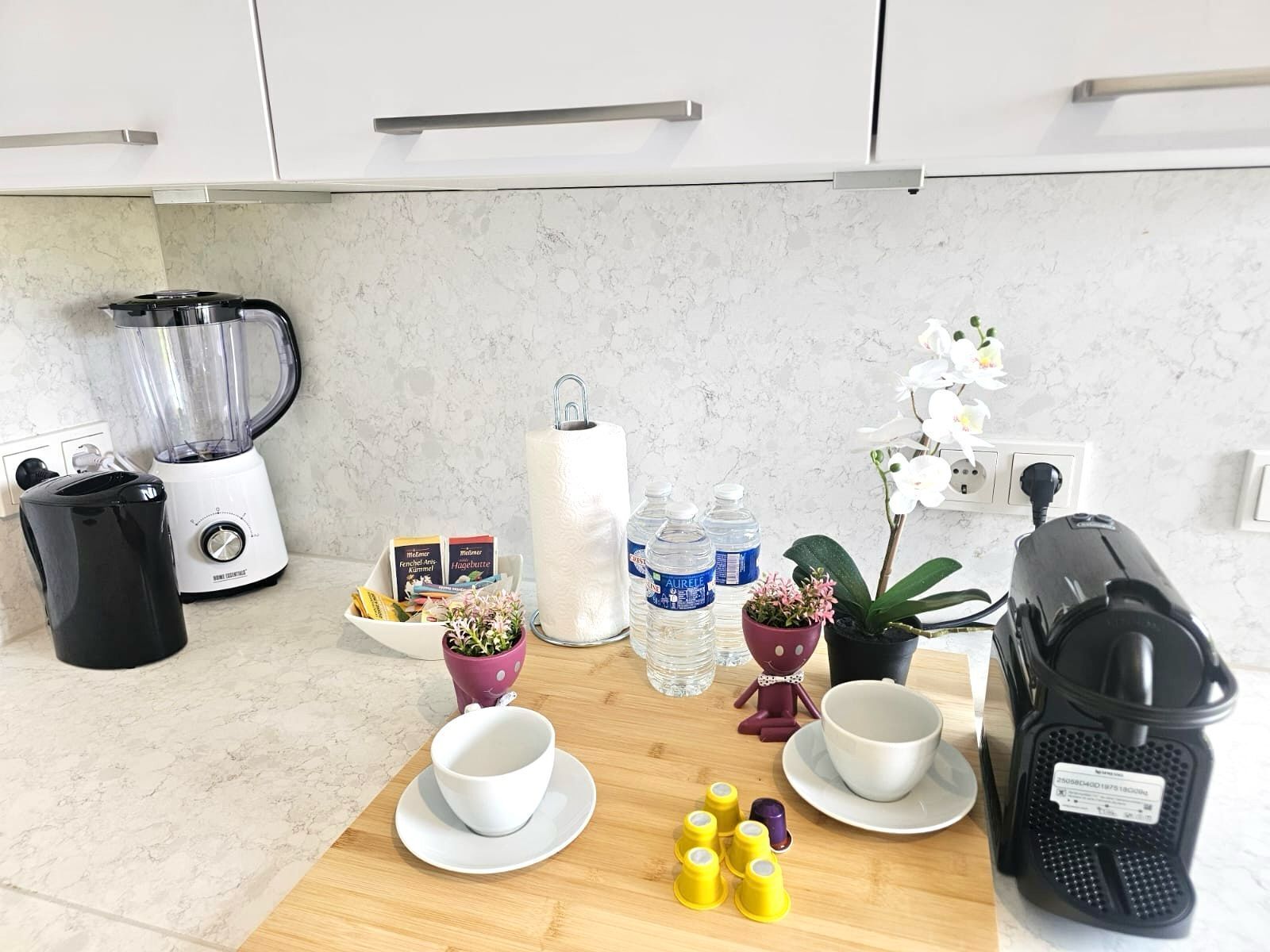 A kitchen with a blender, coffee maker, and a wooden cutting board with cups and utensils.