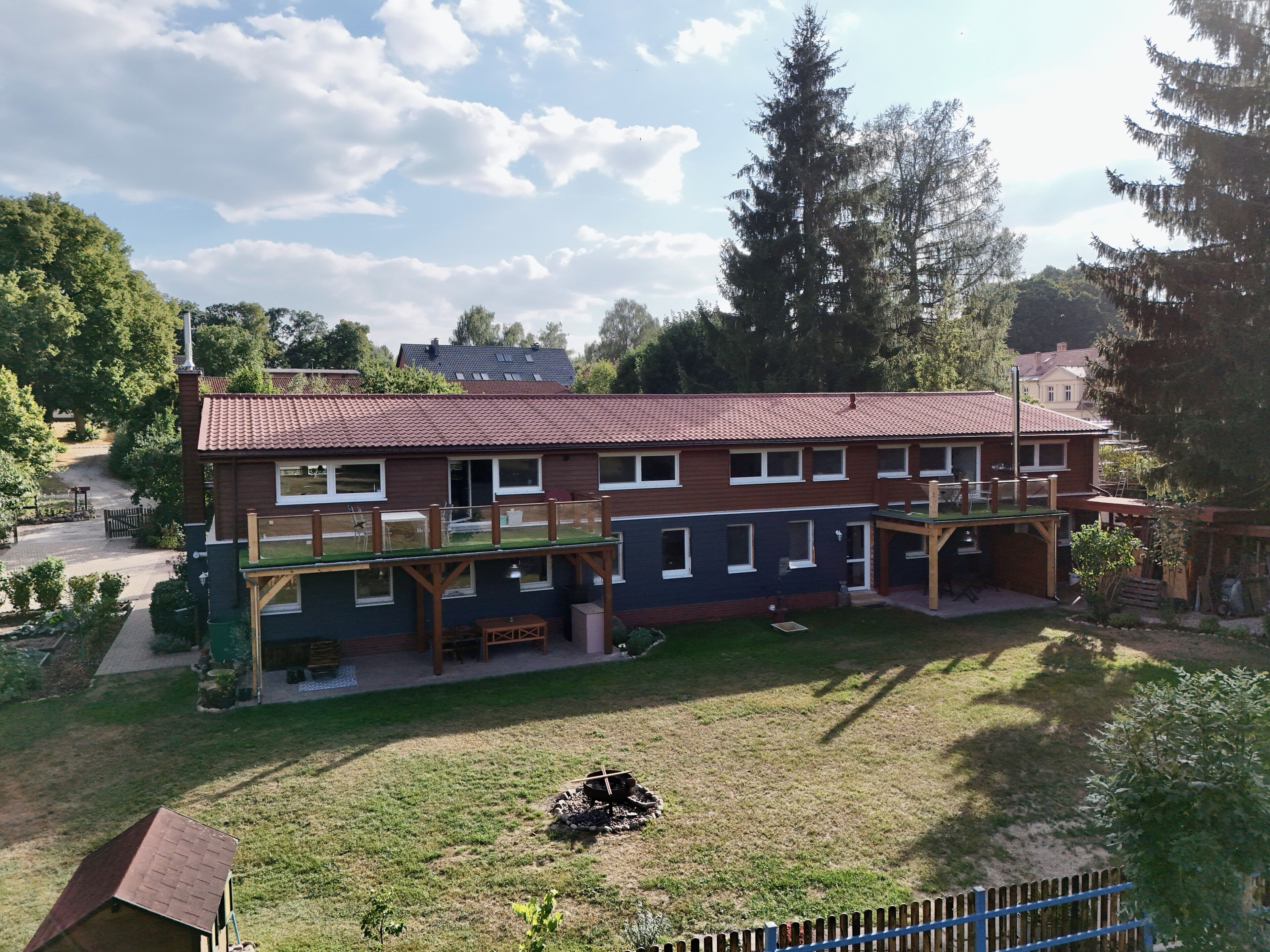 Ein zweistöckiges Gebäude mit rotem Ziegeldach, einer überdachten Terrasse und einem umzäunten Garten.