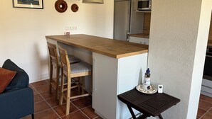 Une cuisine compacte avec un comptoir en bois, un four à micro-ondes encastré et un réfrigérateur. On y trouve une petite table agrémentée d’objets décoratifs et une chaise avec un coussin.