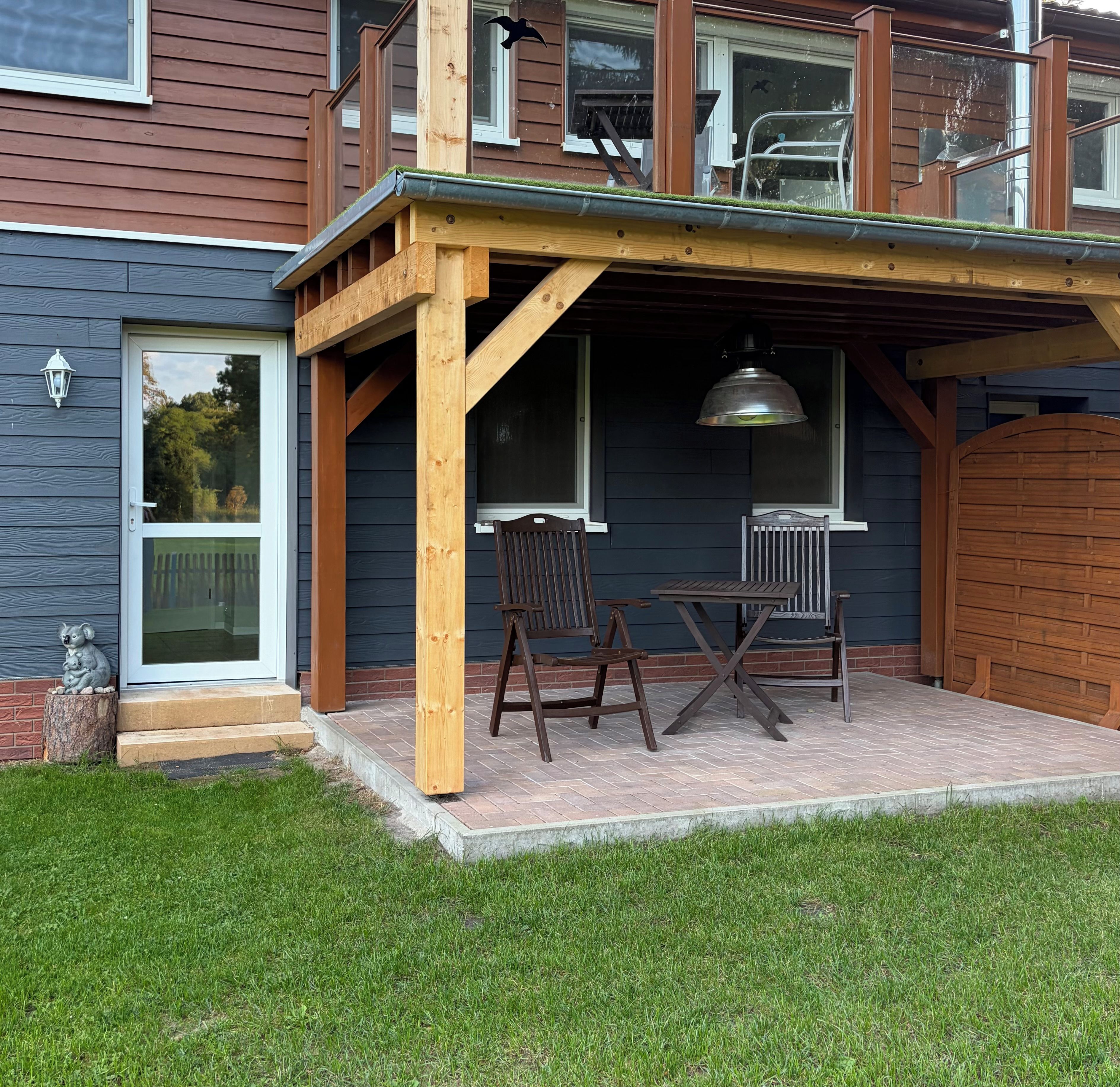 Terraza de madera con mesa y sillas, lámpara colgante y vista de la casa.