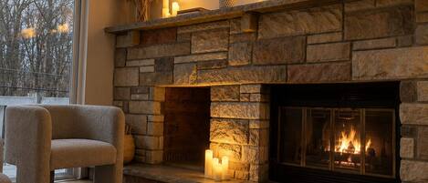 A cozy living room with a stone fireplace, a comfortable armchair, and a large window.