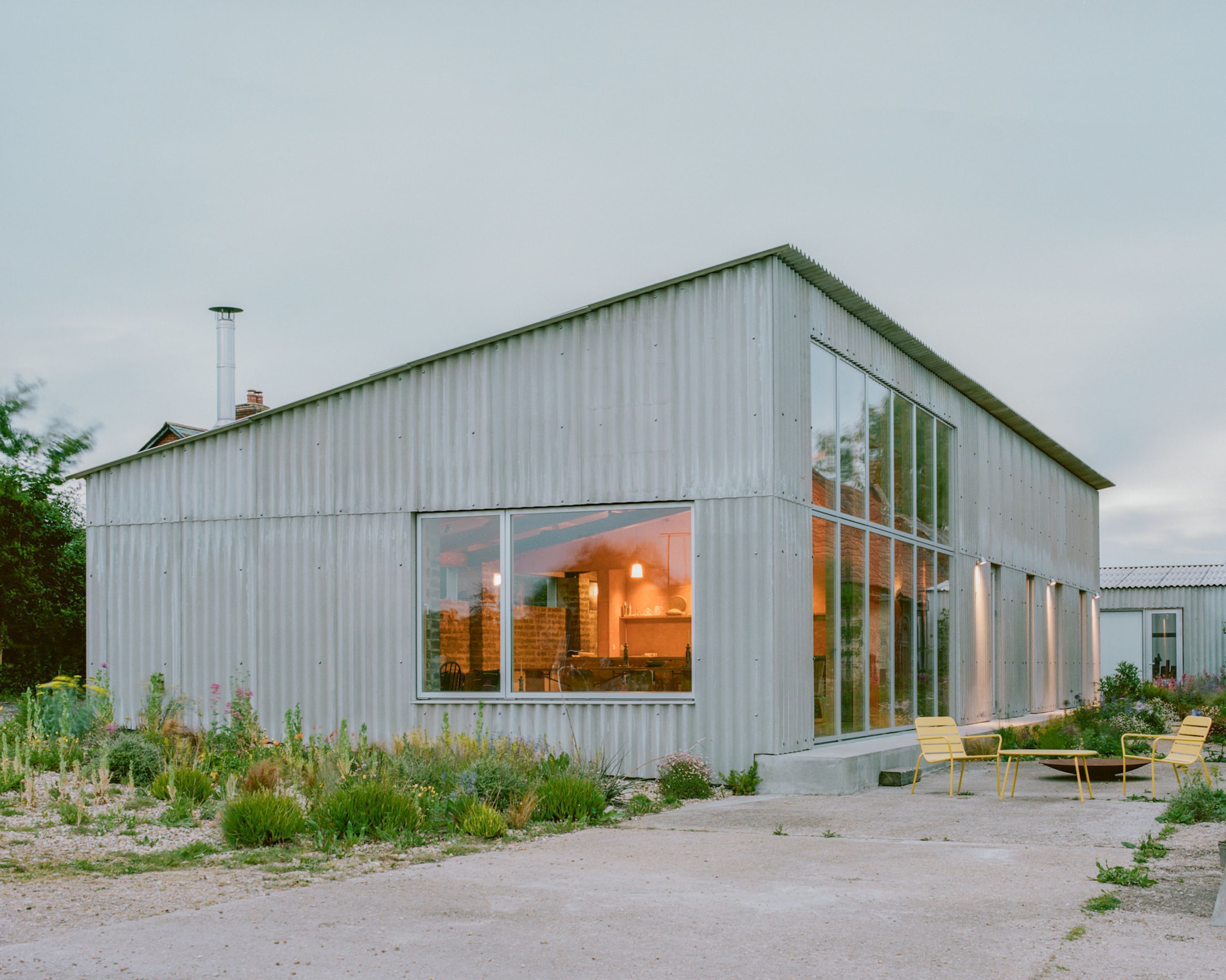 A modern building with large windows, a visible interior with warm lighting, and outdoor seating.