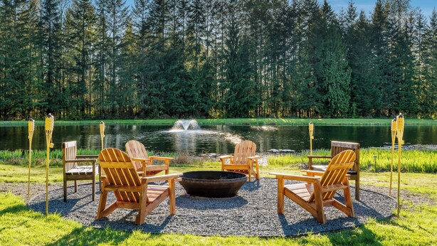 A lakeside seating area with Adirondack chairs and a fire pit, surrounded by a forest and a pond.