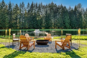 A lakeside seating area with Adirondack chairs and a fire pit, surrounded by a forest and a pond.