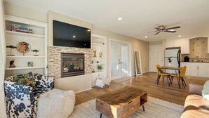 A modern living room with a fireplace, a flat-screen TV, a dining area with yellow chairs, and a kitchen with white cabinets.