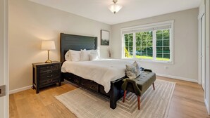 A bedroom with a large bed, a nightstand, a lamp, a bench, and a window with a view of greenery.