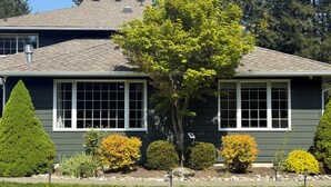 A two-story house with a dark grey exterior, white window frames, and a well-maintained front yard with various shrubs and a tree.