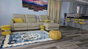 Een moderne woonkamer met een beige bank, gele kussens, een vloerkleed met patroon, en een keukenhoek met barkrukken.