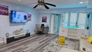 Een moderne woonkamer met een flatscreen televisie, een plafondventilator, een bureau en een zithoek met een bank en een poef.