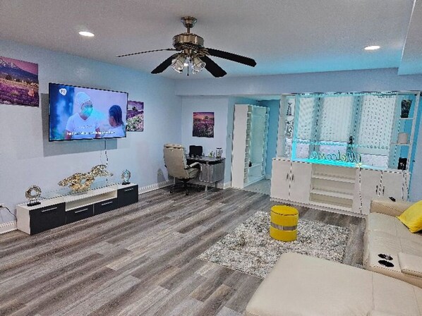 A modern living room with a flat-screen TV, a ceiling fan, a desk, and a seating area with a sofa and ottoman. - Charming 2-bed Garden condo in beautiful Chicago + WiFi, AC, & private parking (Chicago)