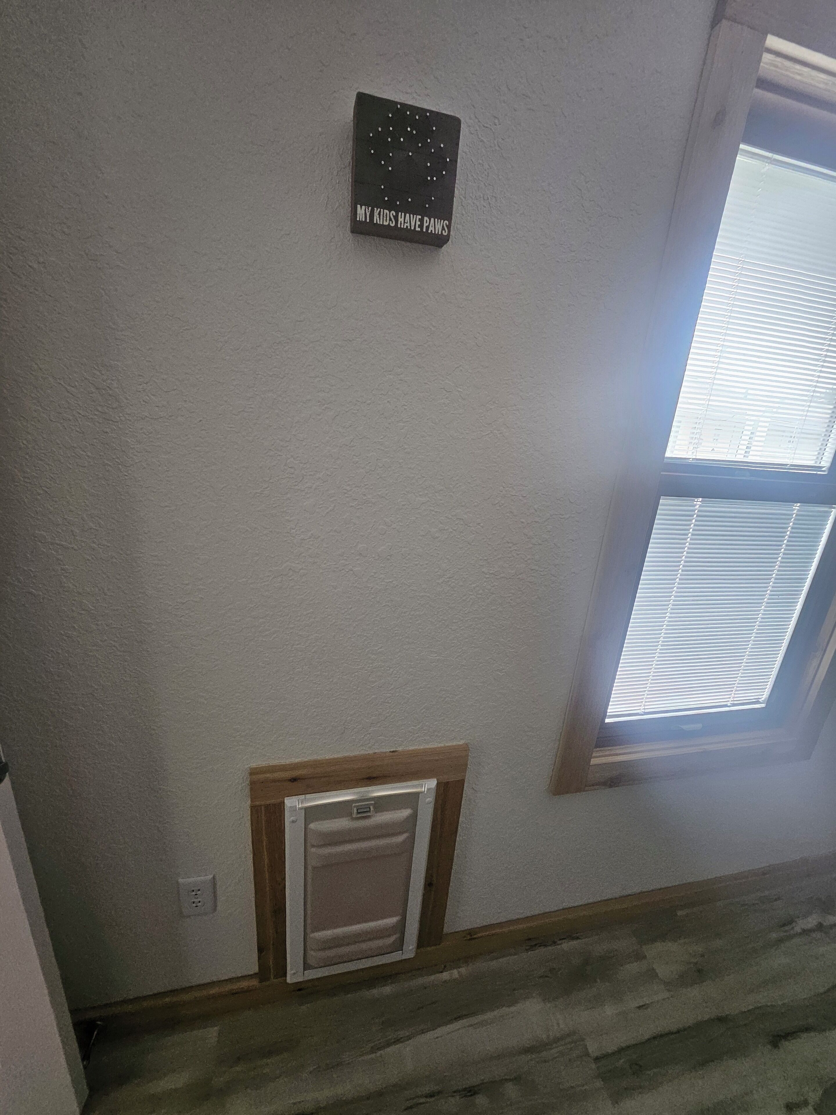 A room with a white wall, a small window with blinds, a thermostat, and a sign that says "MY KIDS HAVE PAWS".