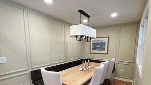 A dining area with a wooden table, white chairs, a black cushioned bench, recessed ceiling lights, and a framed picture on the wall.