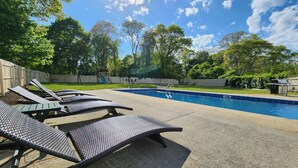 Een achtertuin met een zwembad, lounge fauteuils en een houten terras.
