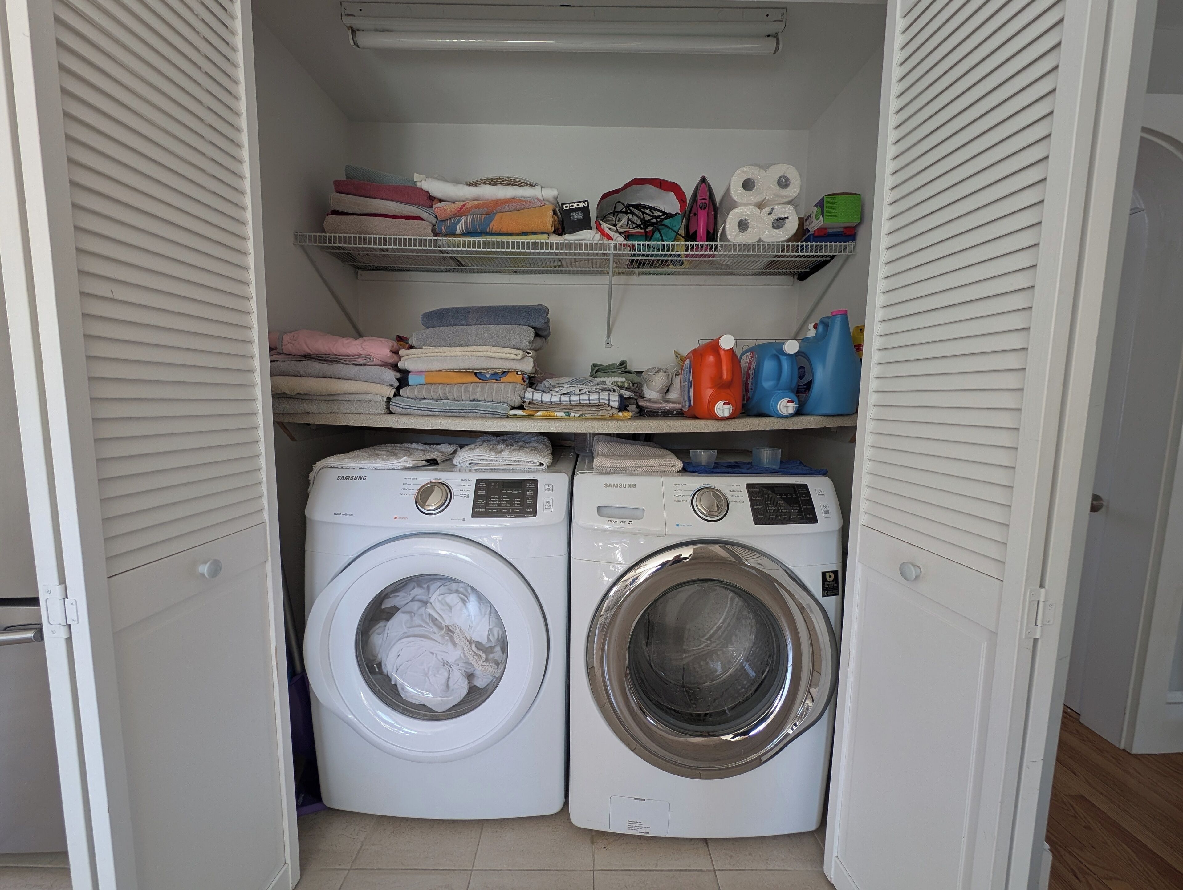 Un cuarto de lavandería con dos lavadoras de carga frontal, repisas superiores con toallas dobladas, una cesta y productos de limpieza.