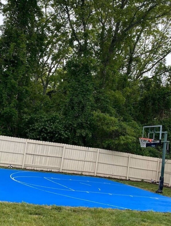 An outdoor basketball court with a blue surface, surrounded by a white fence and trees.