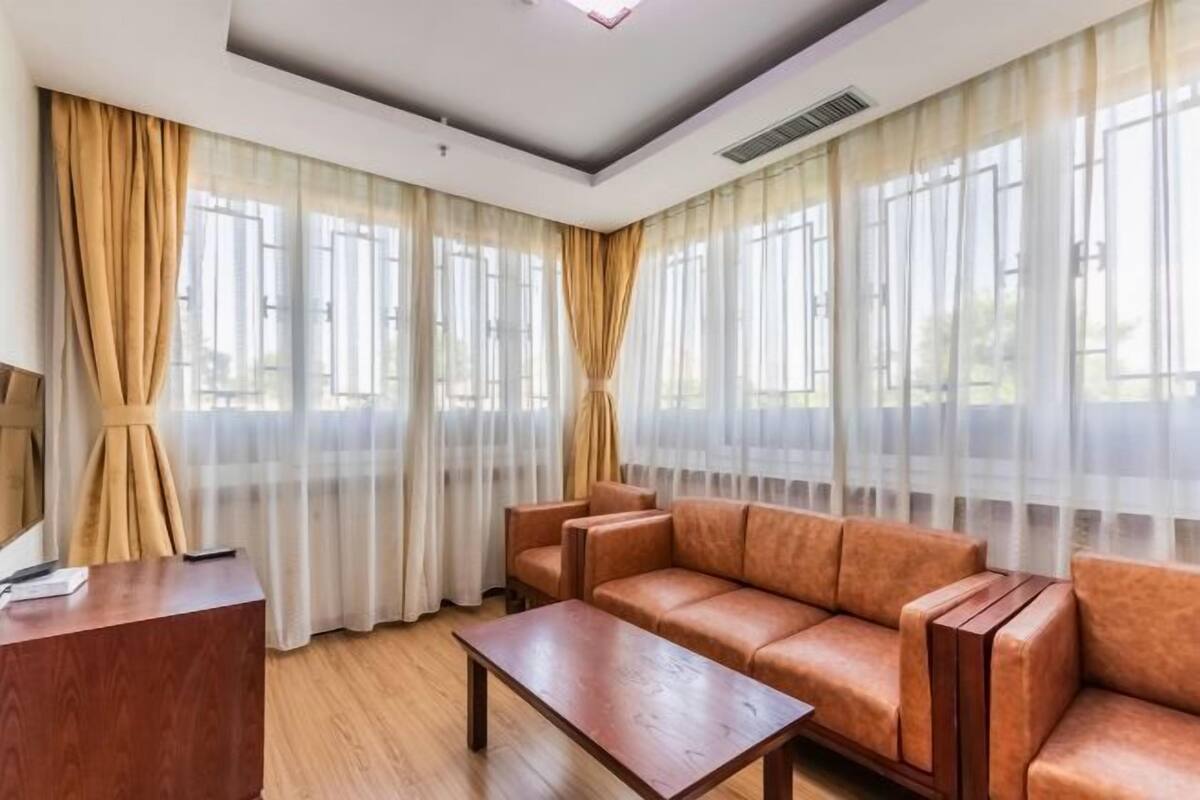Una habitación de hotel moderna con un conjunto de sofás, una mesa de centro de madera y ventanas amplias con cortinas translúcidas.