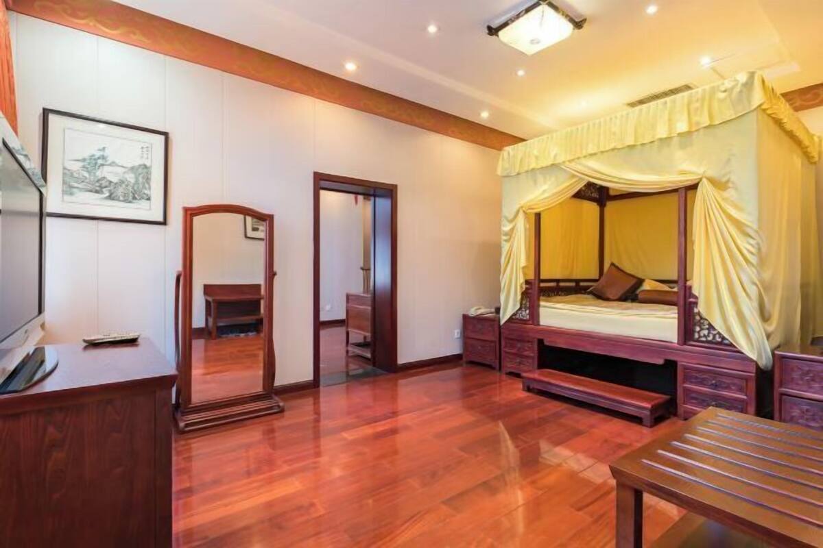 Una habitación con piso de madera, una cama con dosel, un escritorio con una silla, un espejo y un cuadro en la pared.