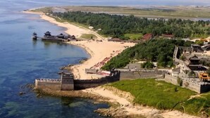 Zona costera con playa, una estructura fortificada y vegetación circundante.