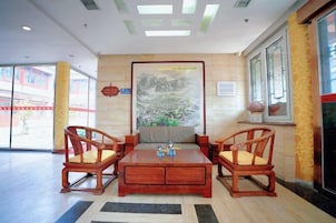 A lobby with a wooden coffee table, two wooden chairs, a large framed painting, and a window with a view.