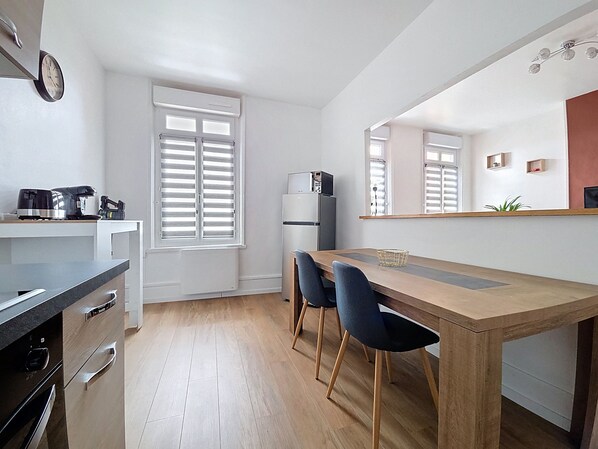 A modern kitchen with a wooden dining table, two chairs, a refrigerator, and a coffee maker.