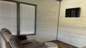Una sala de estar con un sofá gris, un otoman redondo y un televisor de pantalla plana montado en una pared de madera.