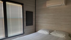 Un dormitorio con cama, ropa de cama blanca, una ventana con persianas y un equipo de aire acondicionado en la pared.