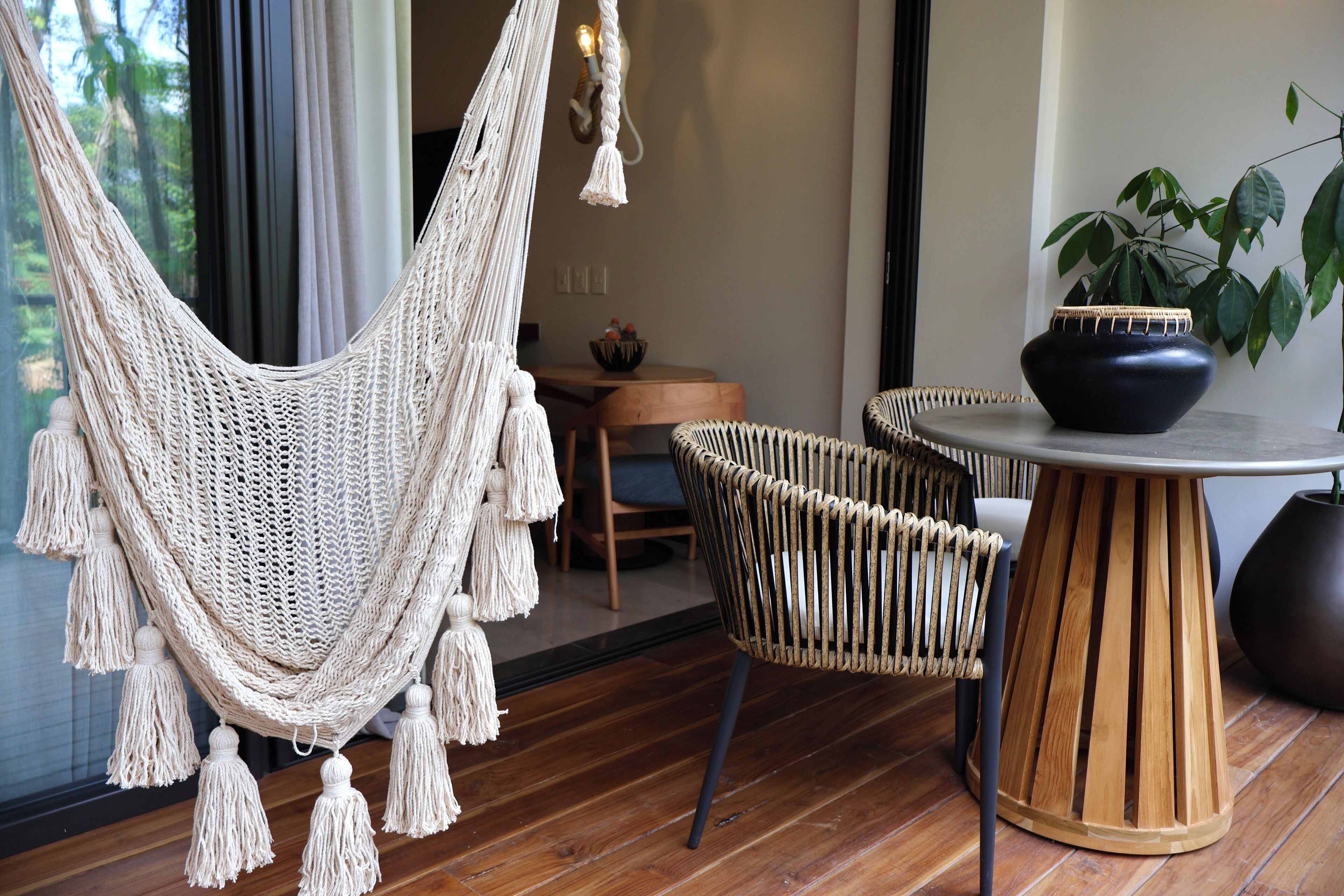 Una habitación con una hamaca de macramé, sillas de mimbre, una mesa redonda de madera y una planta en maceta.