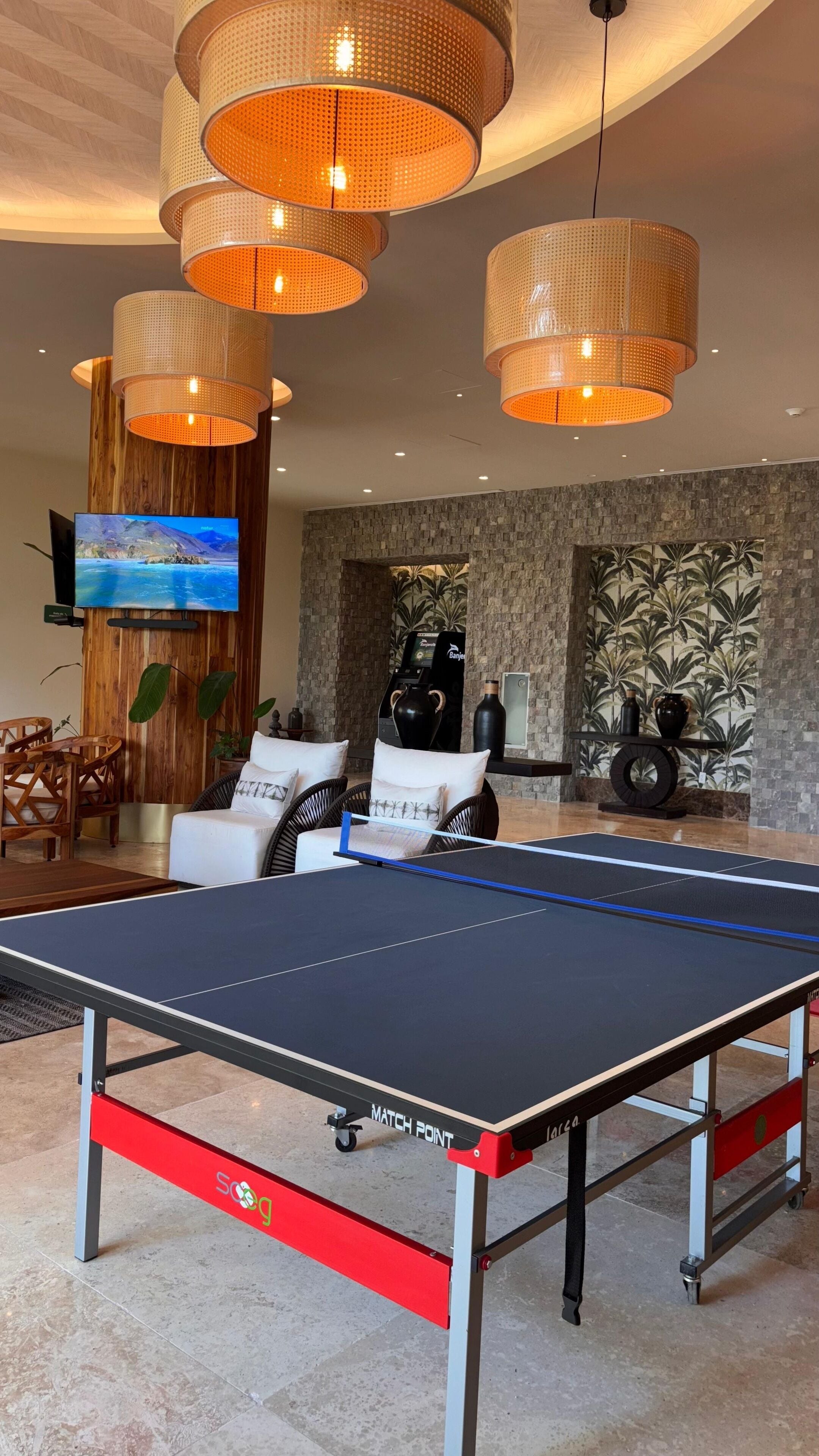 A ping pong table in a modern living room with wicker pendant lights, a flat-screen TV, and a stone wall with a tropical mural.