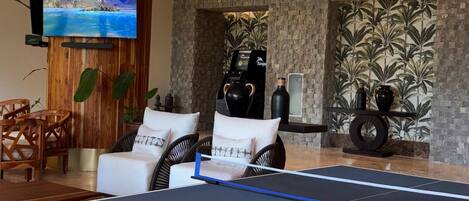 A ping pong table in a modern living room with wicker pendant lights, a flat-screen TV, and a stone wall with a tropical mural.