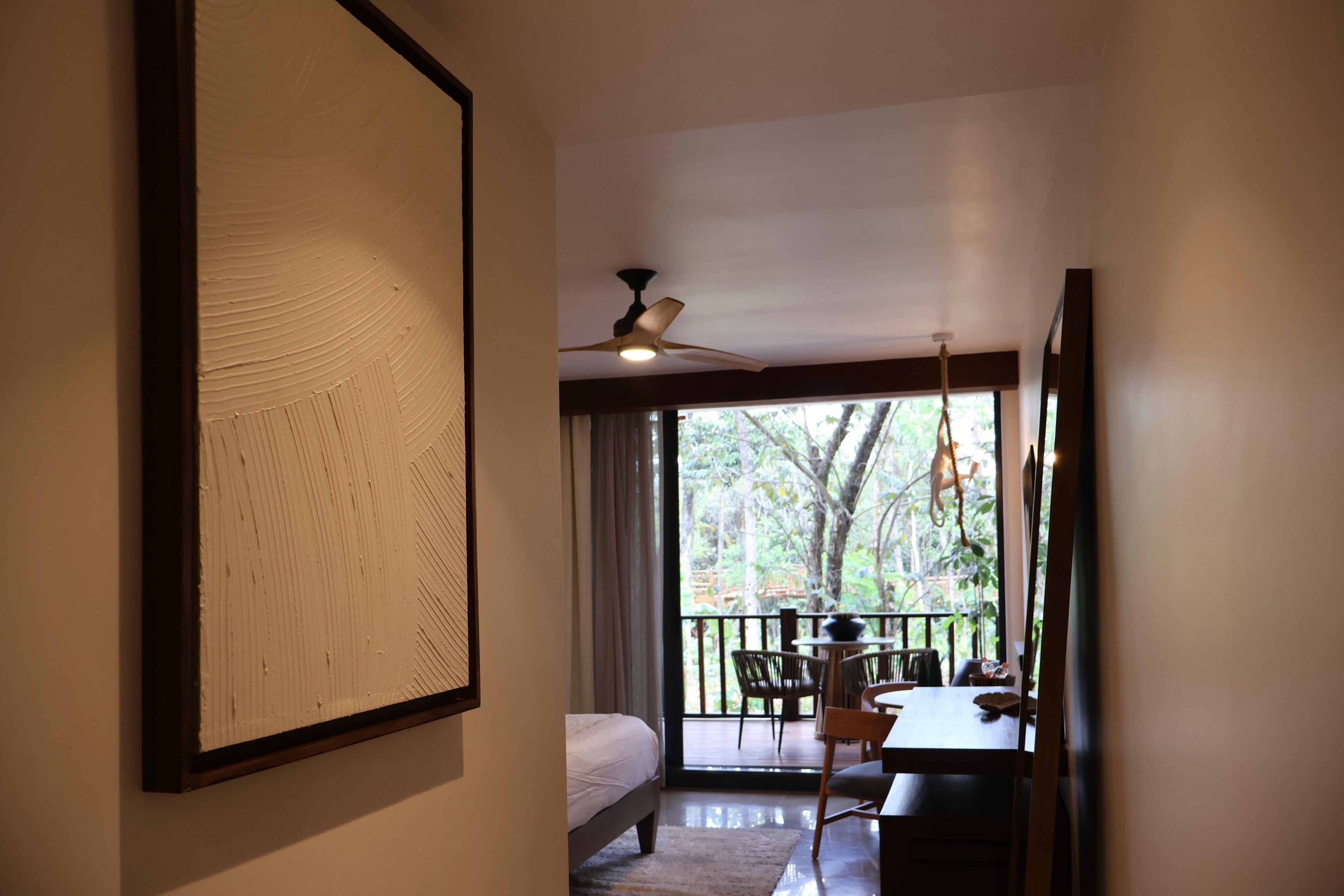 A room with a bed, a dining table, chairs, a ceiling fan, and a view of trees through large windows.